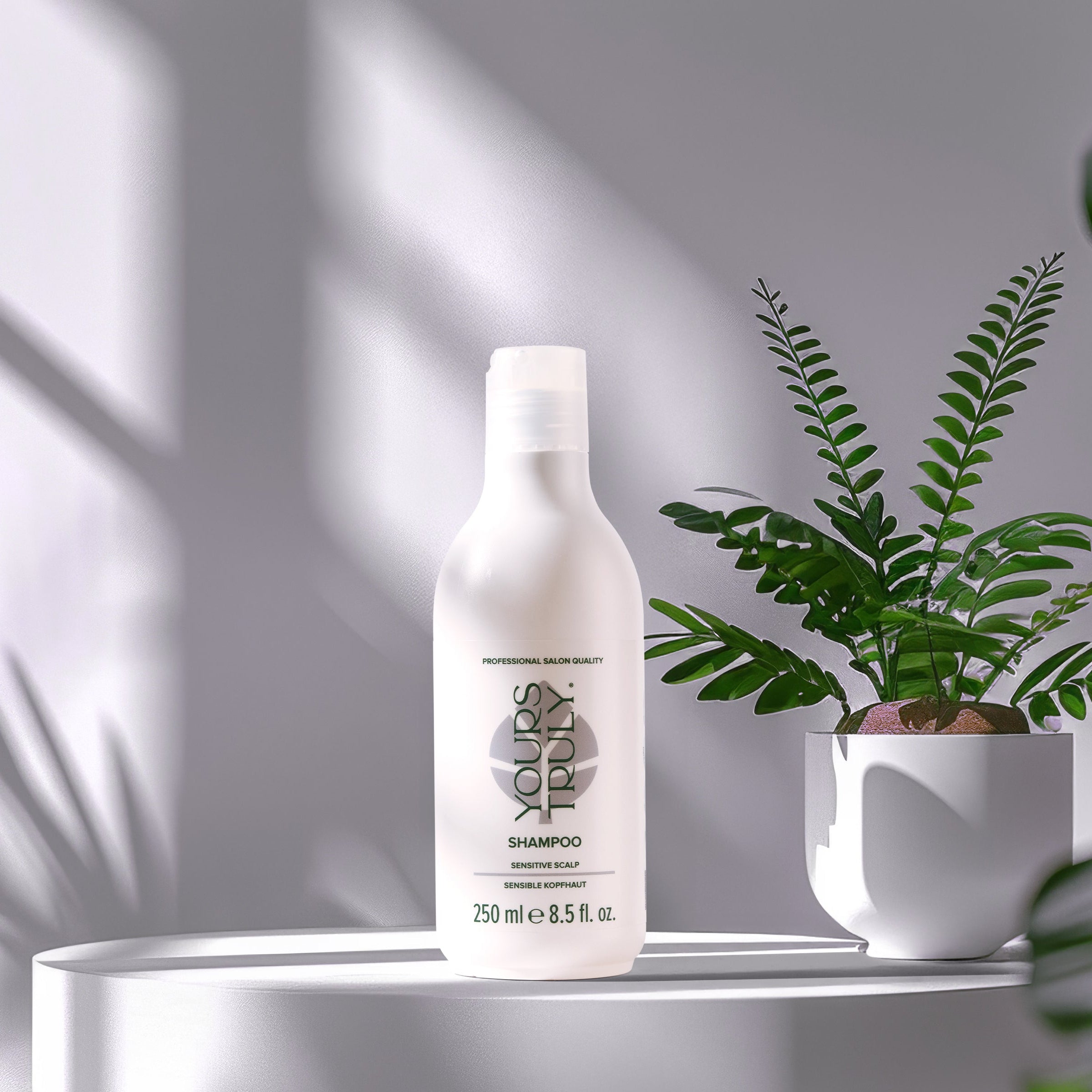 yours-truly-sensitive-scalp-shampoo-in-weißer-250ml-flasche-auf-rundem-podest-mit-grauen-hintergrund-und-topfpflanze