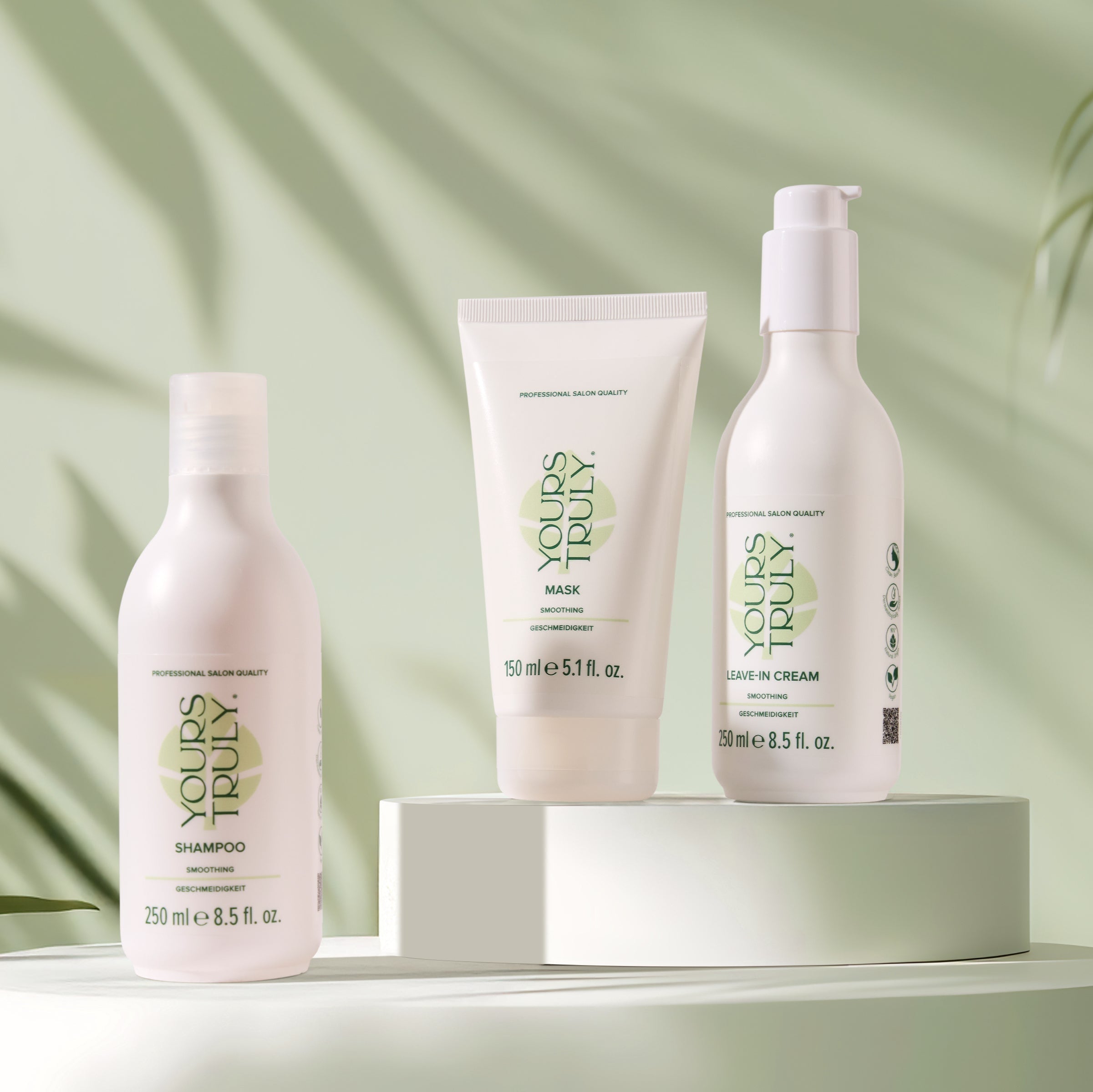 yours-truly-smoothing-shampoo-maske-und-leave-in-cream-in-weißen-gebinden-auf-podest-mit-grünem-hintergrund-und-pflanzenschatten