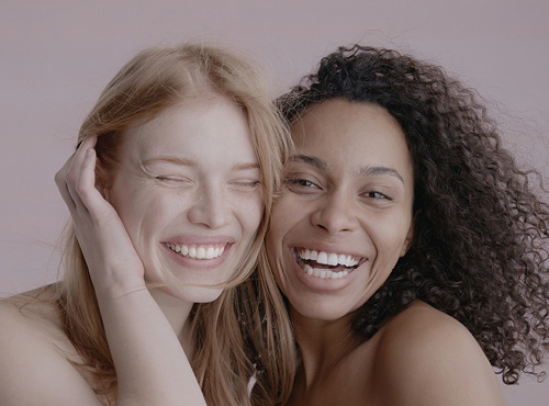 2 Gesichter von Frauen links blond mit glatten Haaren rechts brünette mit braunen haaren beide lachen